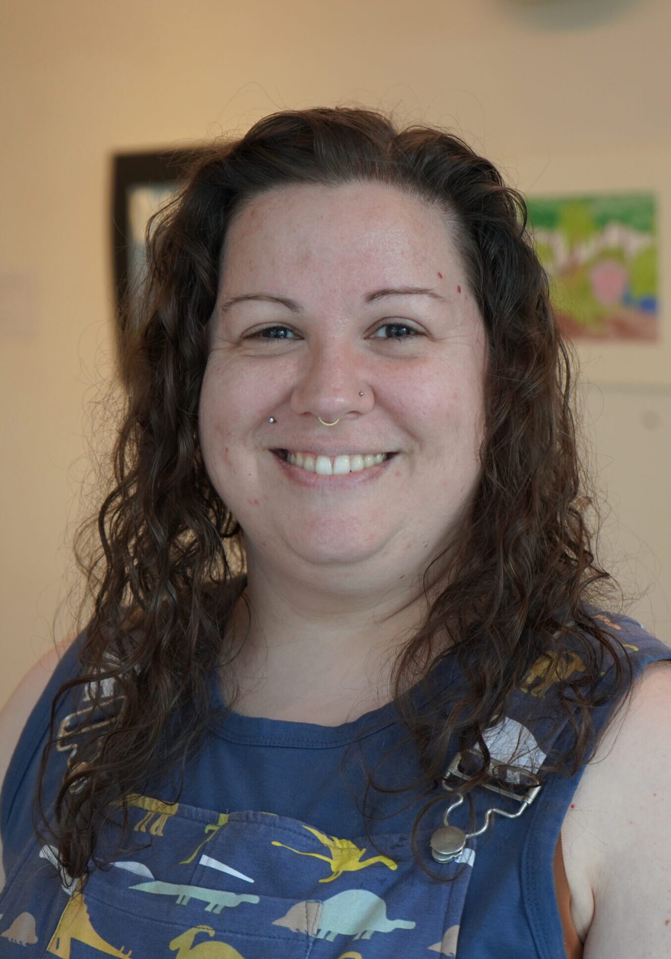 A smiling woman with long, wavy brown hair wears blue overalls patterned with colorful animals while standing in an art gallery.